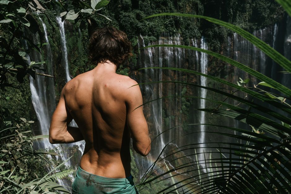 Back view of a shirtless man standing near a waterfall in a tropical forest.